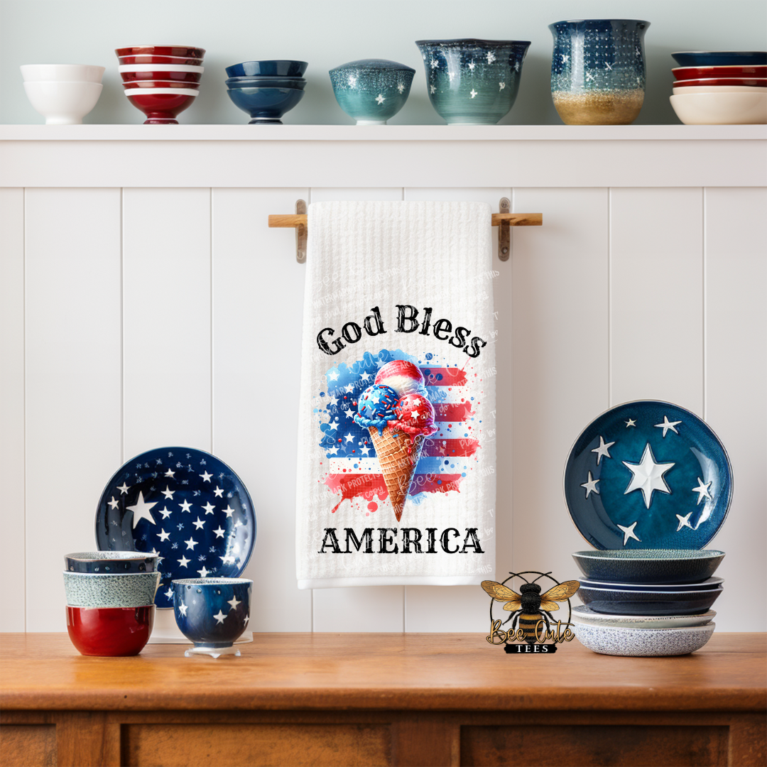White kitchen towel hanging on a wall hook, featuring a patriotic design with a red, white, and blue ice cream cone in front of an American flag and the phrase “God Bless America,” styled in a kitchen with star-themed dishes and bowls on display.
