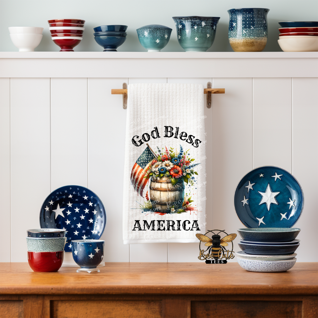 White kitchen towel hanging on a wall hook, featuring a rustic patriotic design with a wooden barrel filled with red, white, and blue flowers and an American flag, along with the phrase “God Bless America,” displayed in a kitchen setting with star-themed dishes and bowls.
