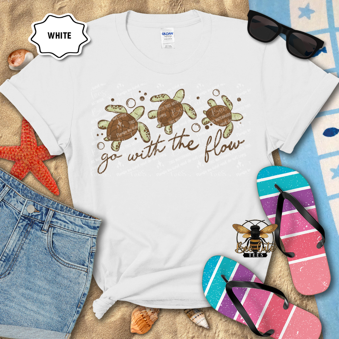 White t-shirt with turtle design and 'go with the flow' text, surrounded by beach items.
