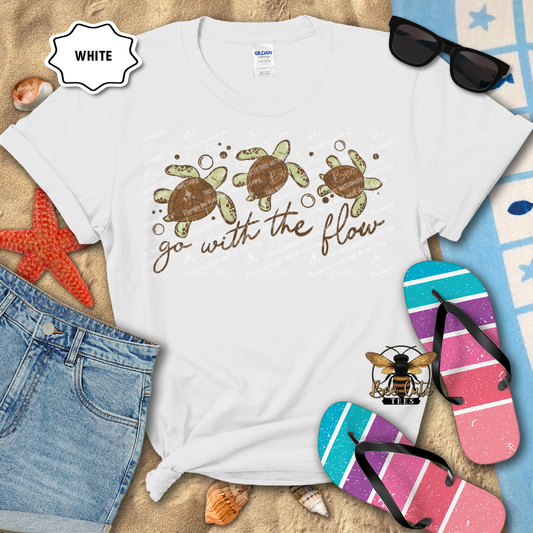 White t-shirt with turtle design and 'go with the flow' text, surrounded by beach items.