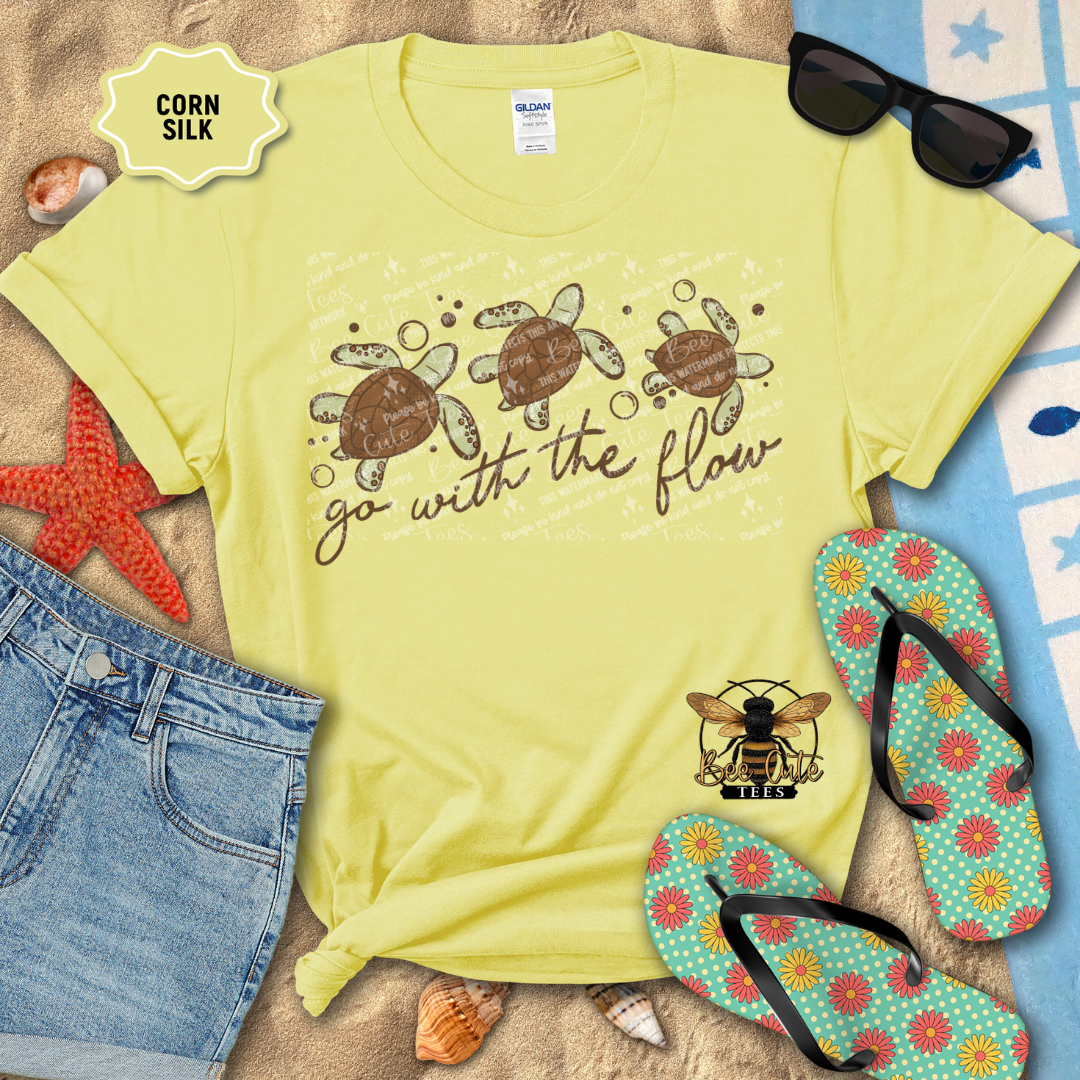 Yellow t-shirt with turtle design and text 'go with the flow' on a beach setting with sunglasses, flip-flops, and starfish.