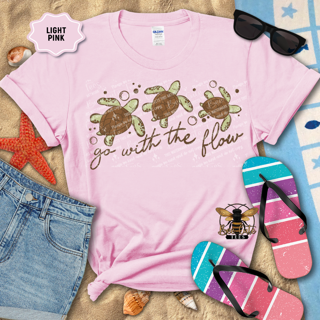 Light pink t-shirt with turtle design and text 'go with the flow' on a beach-themed background.