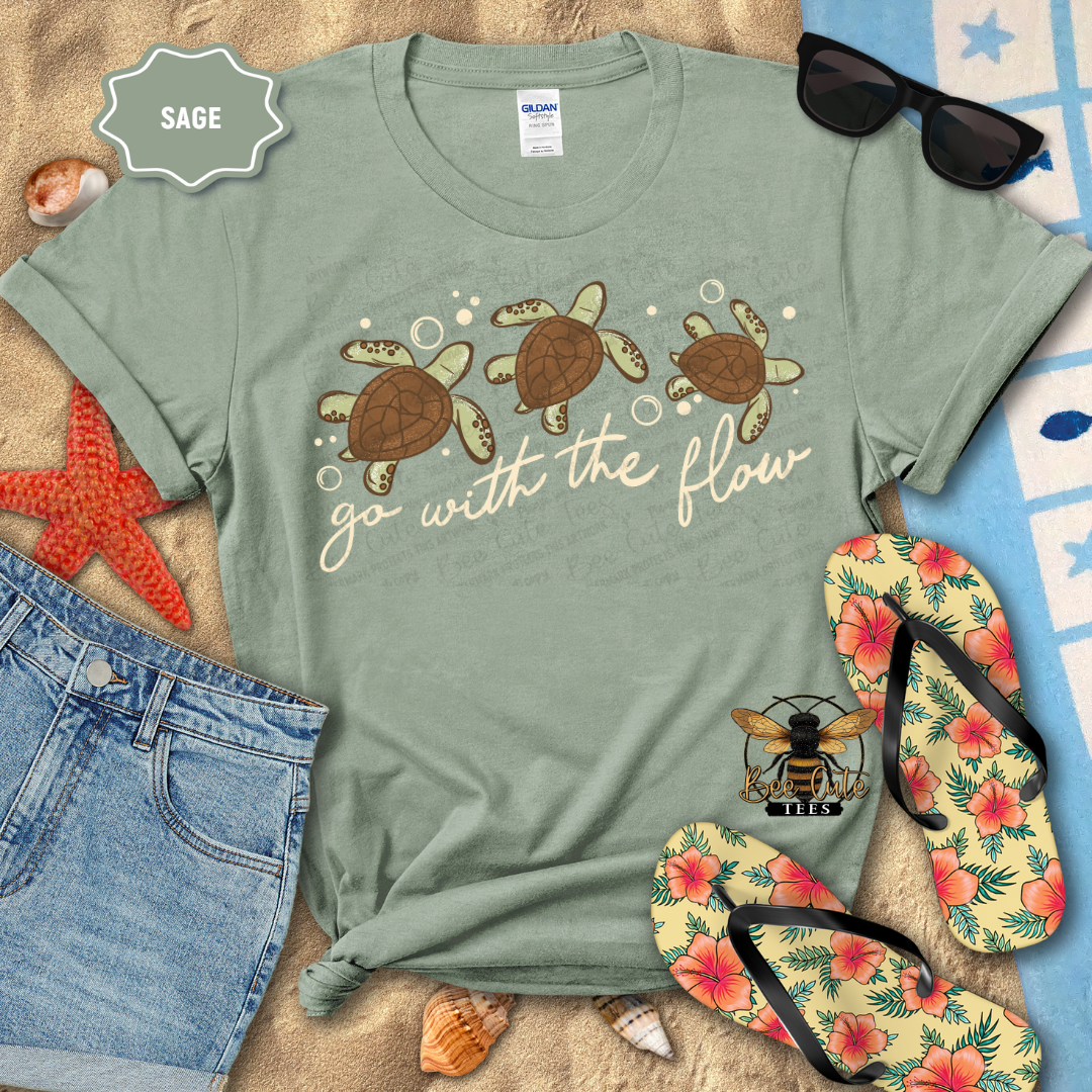 Sage green t-shirt with turtle design and 'go with the flow' text, surrounded by beach accessories.