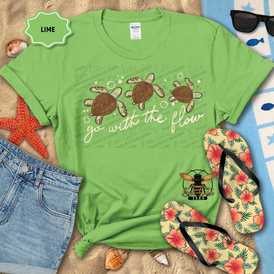 Green t-shirt with turtle design and 'go with the flow' text, surrounded by beach accessories on sand.