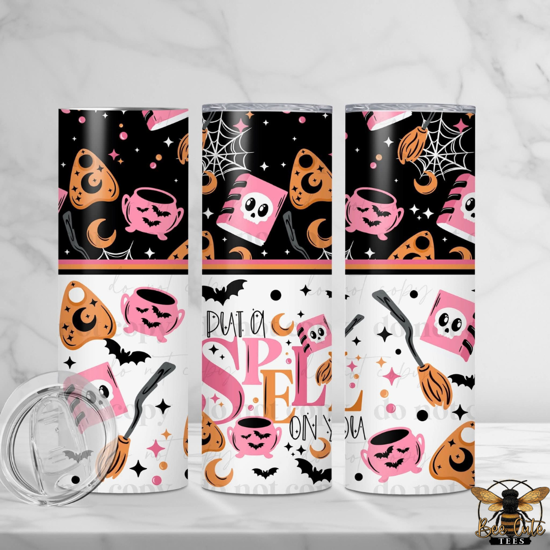 Three Halloween-themed skinny tumblers with a clear lid are displayed on a white marble surface. The tumblers feature a black and white patterned design with pink, orange, and black cartoon cookies, cauldrons, bats, and the text 'PUT A SPELL ON YOU'.