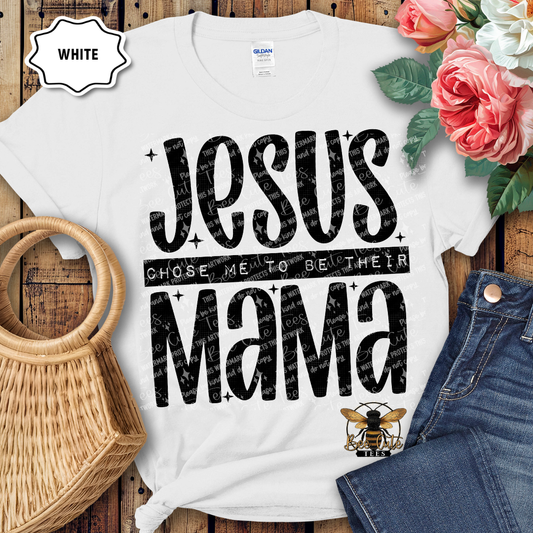 White t-shirt with 'Jesus chose me to be their mama' text on a wooden surface with flowers and denim jeans.