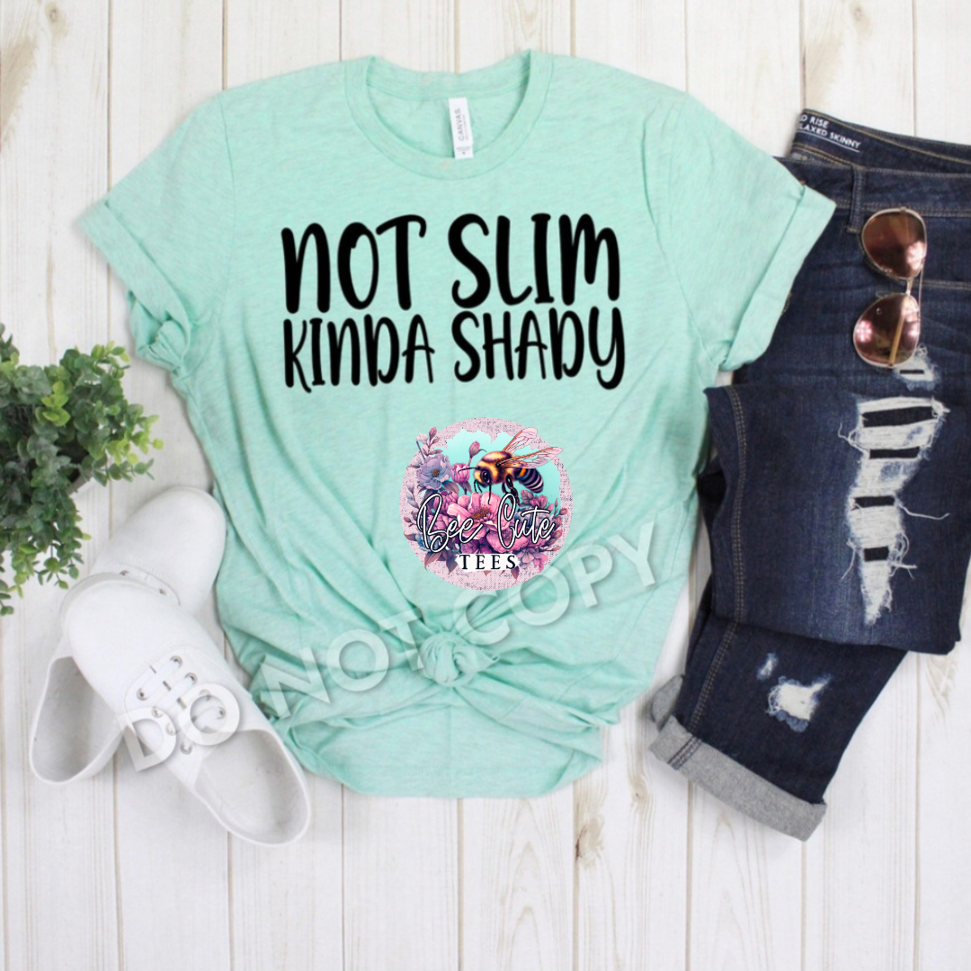Mint green t-shirt with 'Not Slim Kinda Shady' text and bee logo, paired with denim pants and sunglasses on a wooden surface.
