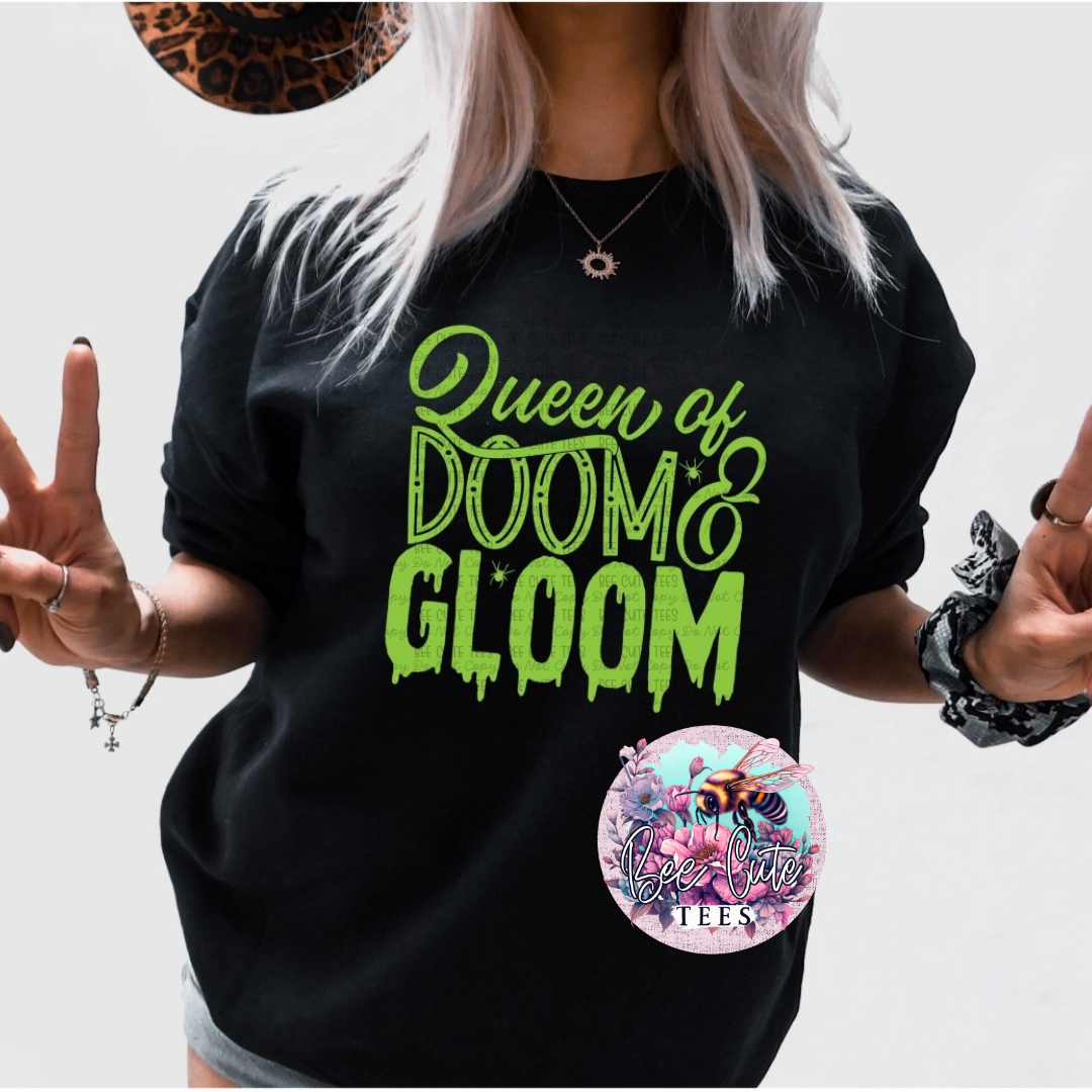 A woman wearing a black graphic t-shirt with the text 'Queen of DOOM & GLOOM' in dripping neon green. She is posing with hands raised, wearing a leopard print hat and jewelry, against a plain white background.