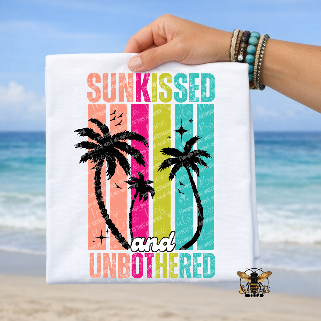 Hand holding a t-shirt with 'Sunkissed and Unbothered' text and palm tree design against a beach background.