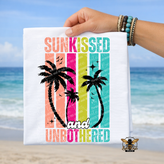Hand holding a t-shirt with 'Sunkissed and Unbothered' text and palm tree design against a beach background.