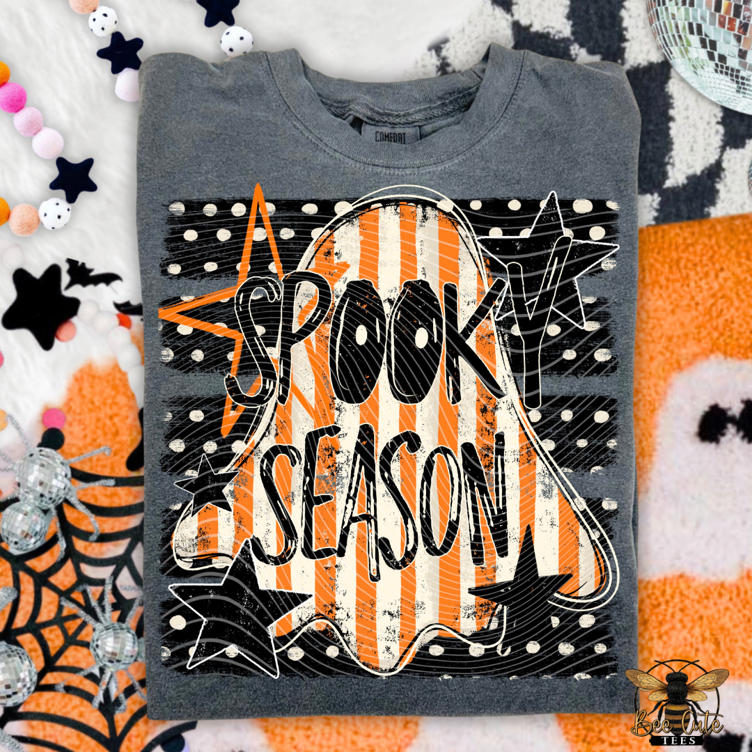 A folded gray crewneck t-shirt with a 'Spooky Season' graphic, styled in a flat lay with Halloween-themed props like a spiderweb, stars, and a disco ball.