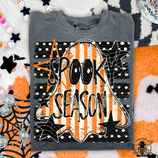 A folded gray crewneck t-shirt with a 'Spooky Season' graphic, styled in a flat lay with Halloween-themed props like a spiderweb, stars, and a disco ball.