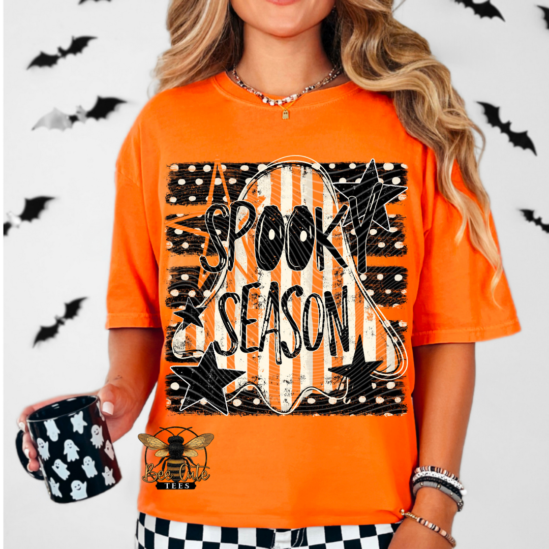 A woman wearing a bright orange graphic t-shirt with 'SPOOKY SEASON' in black and white. She is holding a black mug with white ghosts, set against a white background decorated with black bats.