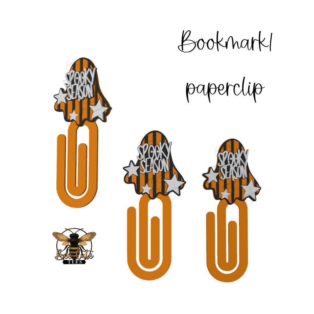 Three orange paperclip bookmarks with a ghost-shaped top featuring orange and black stripes, white stars, and the words 'SPOOKY SEASON,' shown on a white background. A small bee logo appears at lower left, and the words 'Bookmark paperclip' appear at upper right.
