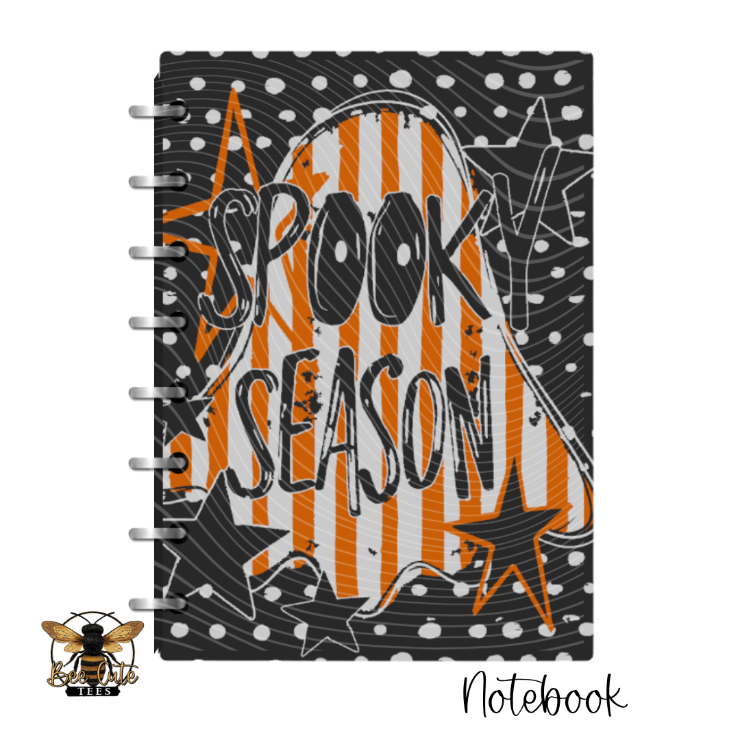 A spiral-bound notebook featuring a Halloween-themed cover design with the words 'SPOOKY SEASON' in black over orange and white ghost stripes, black stars, white dots, and a black background. A small bee logo appears in the lower-left corner, and 'Notebook' is written in script at the lower-right.
