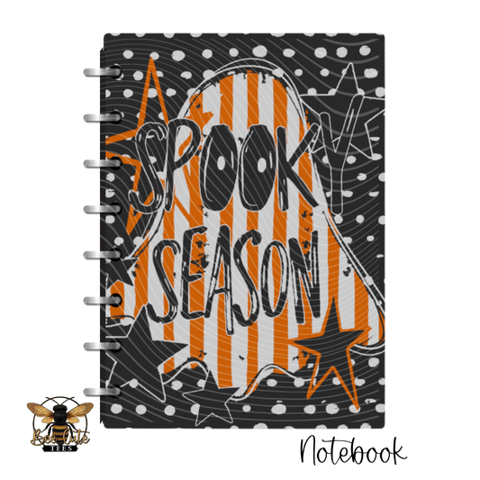 A spiral-bound notebook featuring a Halloween-themed cover design with the words 'SPOOKY SEASON' in black over orange and white ghost stripes, black stars, white dots, and a black background. A small bee logo appears in the lower-left corner, and 'Notebook' is written in script at the lower-right.