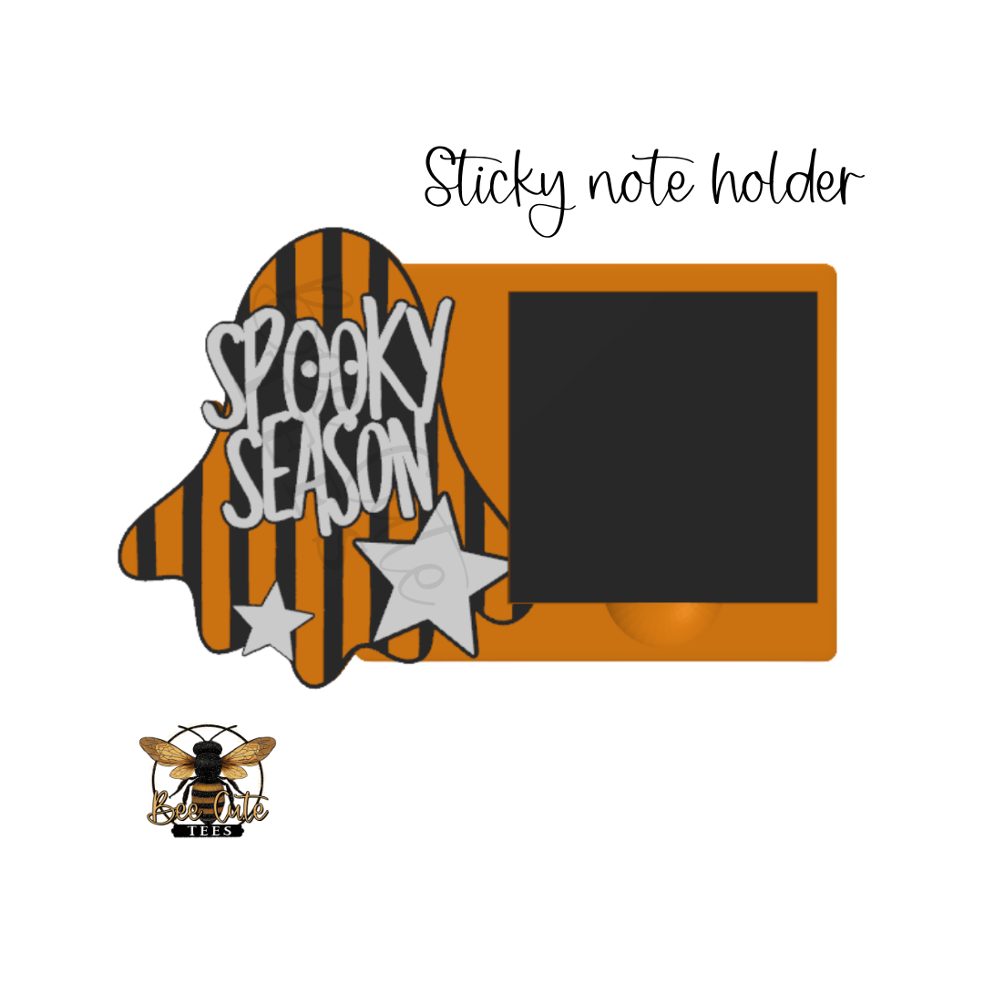 A Halloween-themed sticky note holder featuring a ghost-shaped section with orange and black stripes, the words 'SPOOKY SEASON' in white, two white stars, and an orange rectangular tray on a white background. Handwritten text above reads 'Sticky note holder,' and a small bee logo appears at the bottom left.