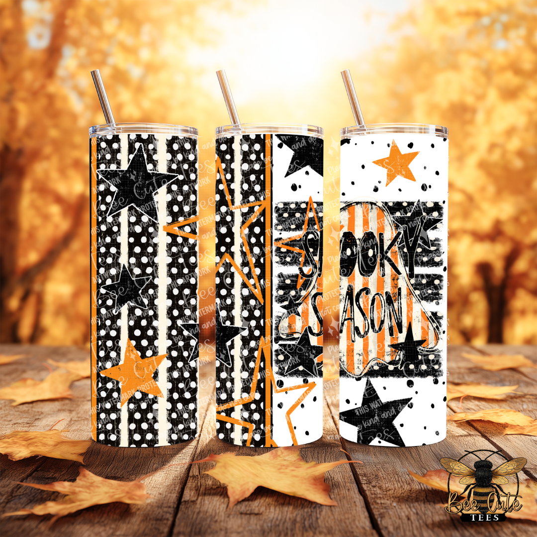 Three insulated tumblers with clear lids and metal straws, decorated with black and orange Halloween-themed graphics including stars, stripes, polka dots, and the words 'SPOOKY SEASON,' set on a wooden surface with autumn leaves and a warm blurred tree background.