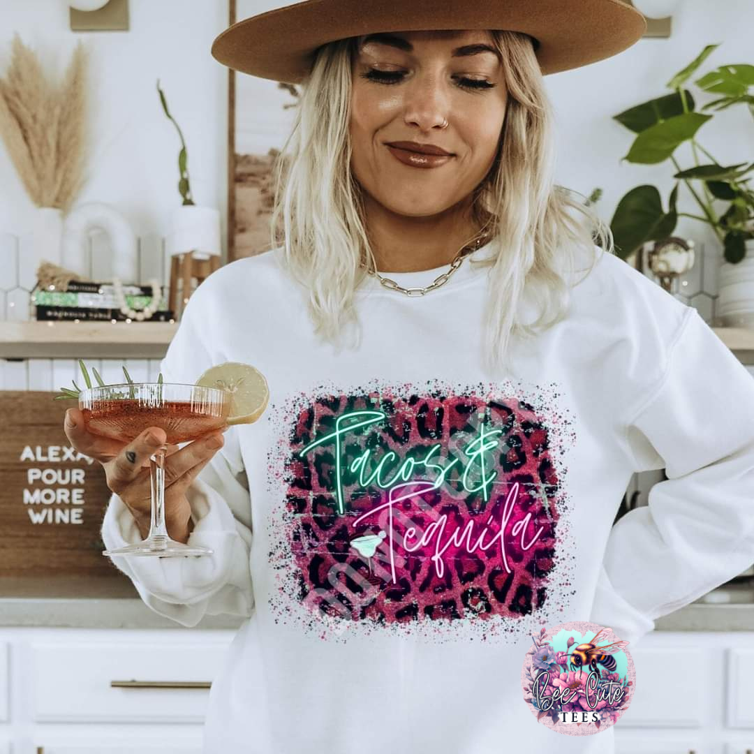 Woman wearing a white sweatshirt with 'Tacos & Tequila' design, holding a drink.