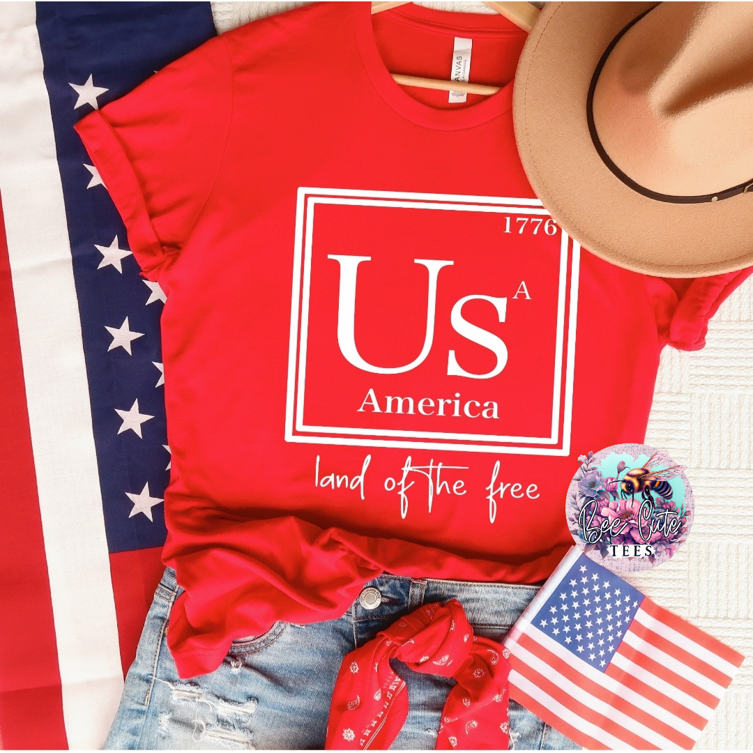 Red graphic t-shirt with a bold “USA America 1776 – Land of the Free” design, styled with distressed denim shorts, a red bandana, a tan hat, and American flag accents for a classic patriotic look.
