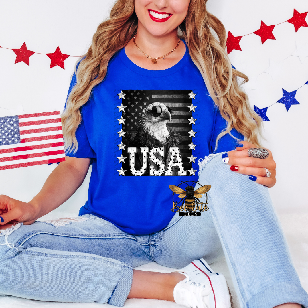 Woman seated in a patriotic-themed setting wearing a royal blue graphic tee featuring a bald eagle in sunglasses, a distressed American flag background, and bold “USA” lettering framed by stars. Styled with light-wash jeans, white sneakers, patriotic nail colors, and holding an American flag, with red and blue star decorations in the background.
