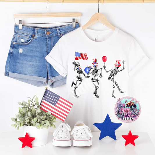 White graphic t-shirt featuring dancing skeletons with patriotic hats, balloons, and an American flag, styled with denim shorts, sneakers, and festive red, white, and blue decor.
