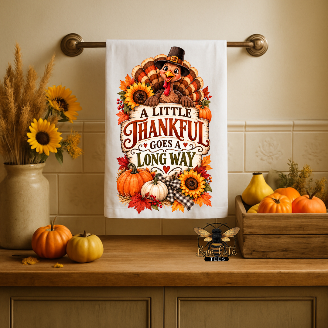 A white kitchen towel with a Thanksgiving-themed turkey and autumn quote hangs on a brass towel bar above a wooden counter. The towel features a cartoon turkey wearing a pilgrim hat, surrounded by sunflowers, pumpkins, and the text 'A LITTLE THANKFUL GOES LONG WAY'.