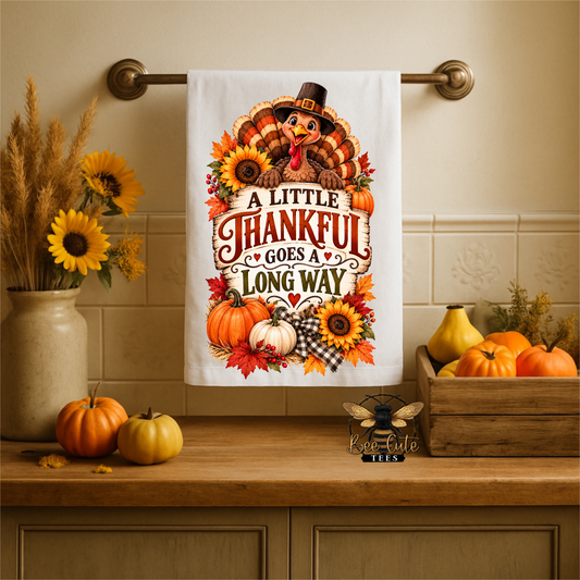 A white kitchen towel with a Thanksgiving-themed turkey and autumn quote hangs on a brass towel bar above a wooden counter. The towel features a cartoon turkey wearing a pilgrim hat, surrounded by sunflowers, pumpkins, and the text 'A LITTLE THANKFUL GOES LONG WAY'.