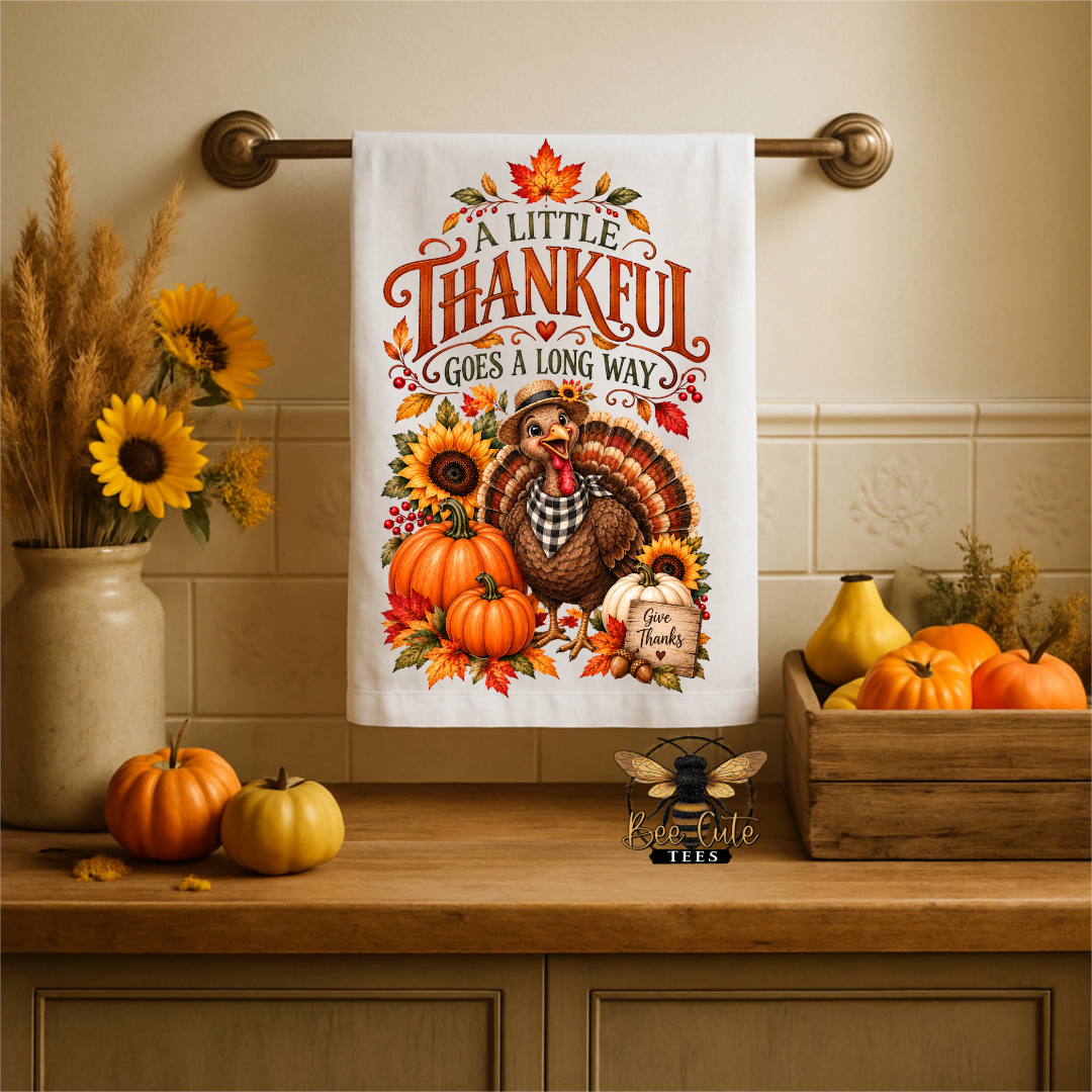 A white kitchen towel with a colorful Thanksgiving-themed illustration hangs on a brass towel bar. The towel features the text 'A LITTLE THANKFUL GOES A LONG WAY' above a turkey, pumpkins, sunflowers, and autumn leaves.