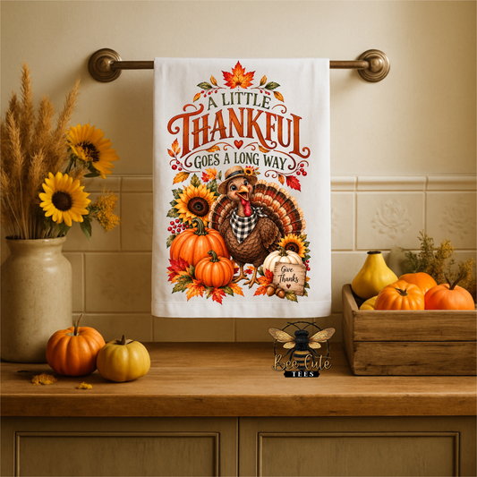 A white kitchen towel with a colorful Thanksgiving-themed illustration hangs on a brass towel bar. The towel features the text 'A LITTLE THANKFUL GOES A LONG WAY' above a turkey, pumpkins, sunflowers, and autumn leaves.