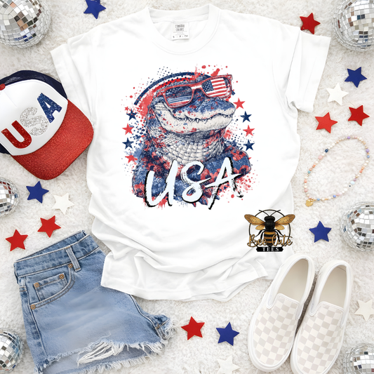 White t-shirt with alligator graphic and 'USA' text, surrounded by accessories on a light background.