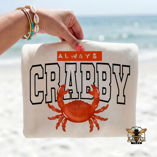 Beige T-shirt with 'Always Crabby' text and a crab illustration held by a hand on a beach.