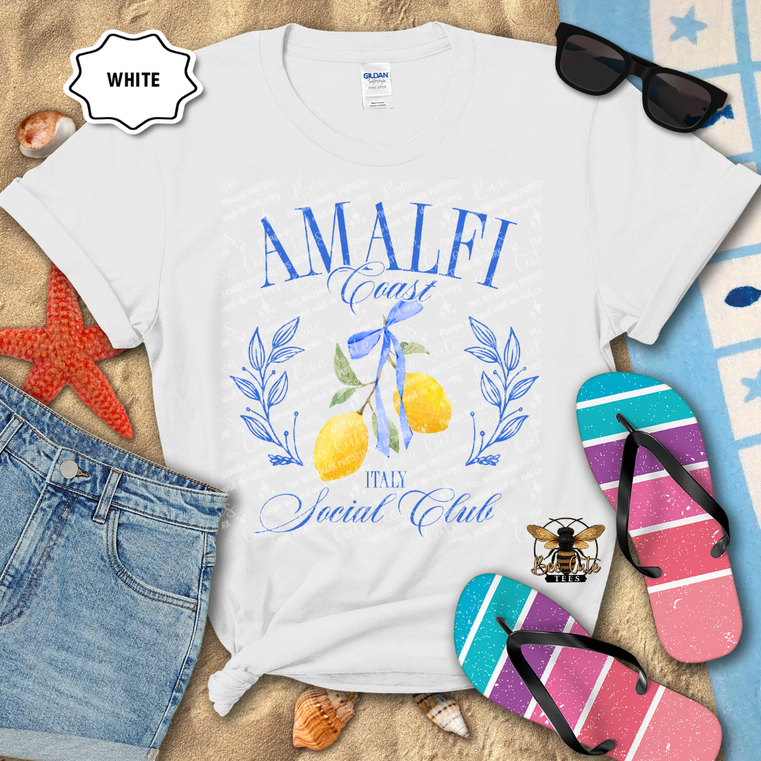 White Gildan Softstyle T-shirt displayed on a beach-themed flat lay with denim shorts, flip-flops, sunglasses, seashells, and a starfish. The shirt features a blue "Amalfi Coast Social Club" design with elegant script lettering, illustrated yellow lemons tied with a blue ribbon, and "Italy" beneath the artwork. The overall design evokes a coastal Italian summer aesthetic with bright blue and yellow accents.
