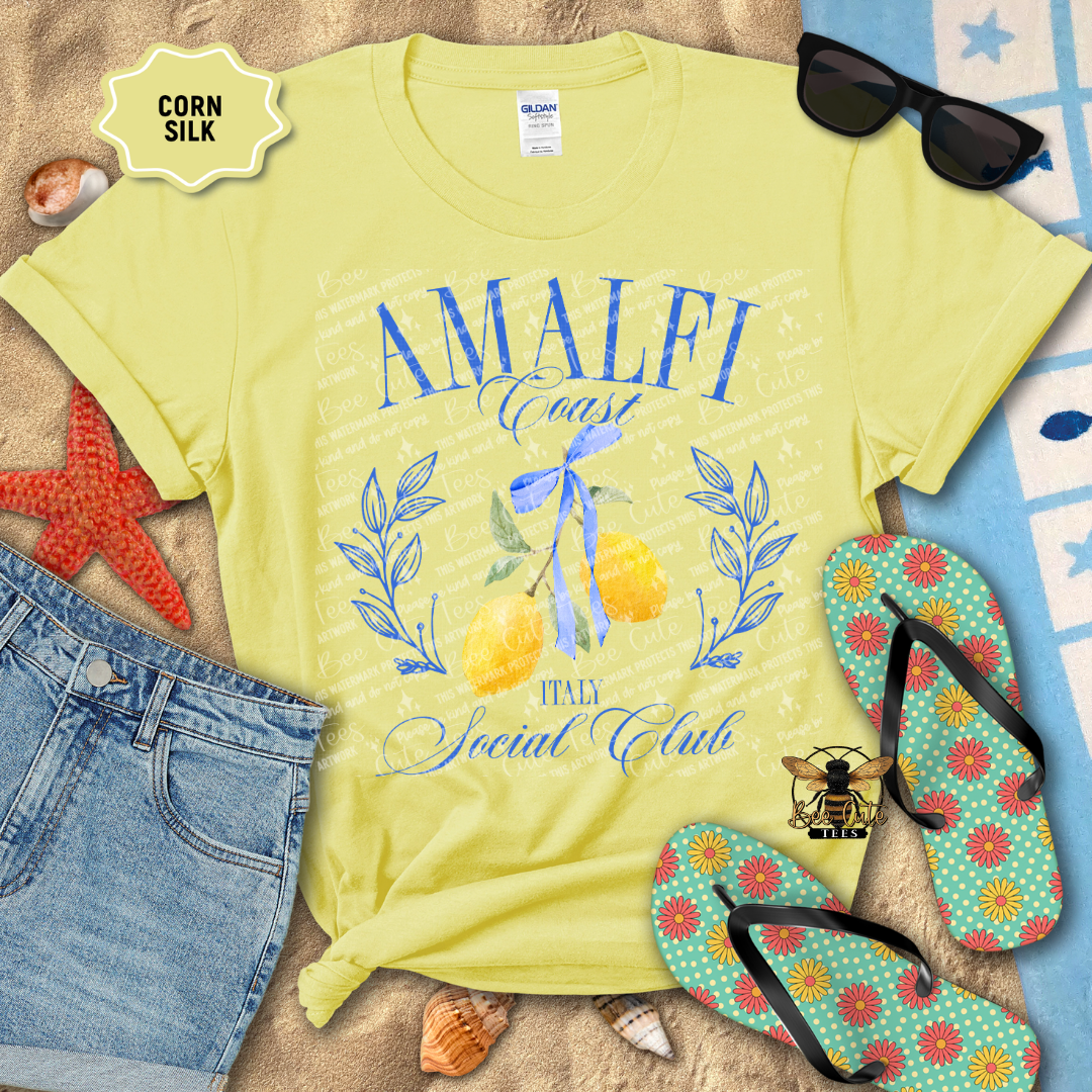 Yellow Gildan Softstyle T-shirt displayed on a beach-themed flat lay with denim shorts, flip-flops, sunglasses, seashells, and a starfish. The shirt features a blue "Amalfi Coast Social Club" design with elegant script lettering, illustrated yellow lemons tied with a blue ribbon, and "Italy" beneath the artwork. The overall design evokes a coastal Italian summer aesthetic with bright blue and yellow accents.
