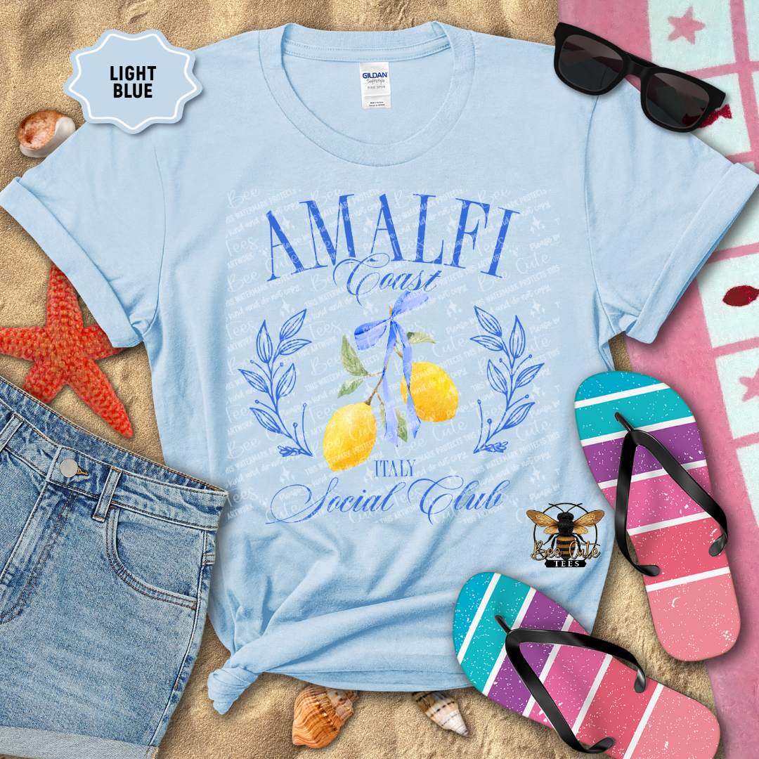 Light Blue Gildan Softstyle T-shirt displayed on a beach-themed flat lay with denim shorts, flip-flops, sunglasses, seashells, and a starfish. The shirt features a blue "Amalfi Coast Social Club" design with elegant script lettering, illustrated yellow lemons tied with a blue ribbon, and "Italy" beneath the artwork. The overall design evokes a coastal Italian summer aesthetic with bright blue and yellow accents.

