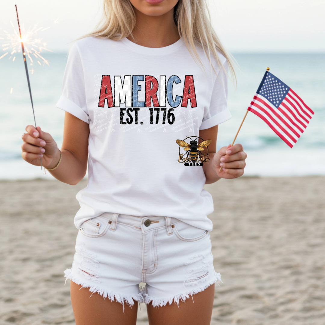 Person wearing a white t-shirt with 'AMERICA EST. 1776' design on a beach.