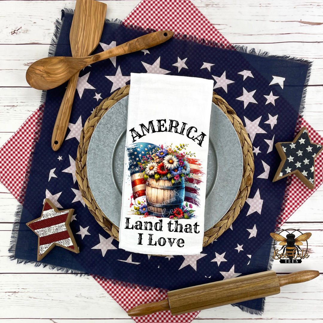 White kitchen towel placed on a patriotic table setting, featuring a wooden barrel filled with red, white, and blue flowers in front of an American flag and the phrase “America, Land that I Love,” surrounded by star-patterned cloths, wooden utensils, and rustic decor.
