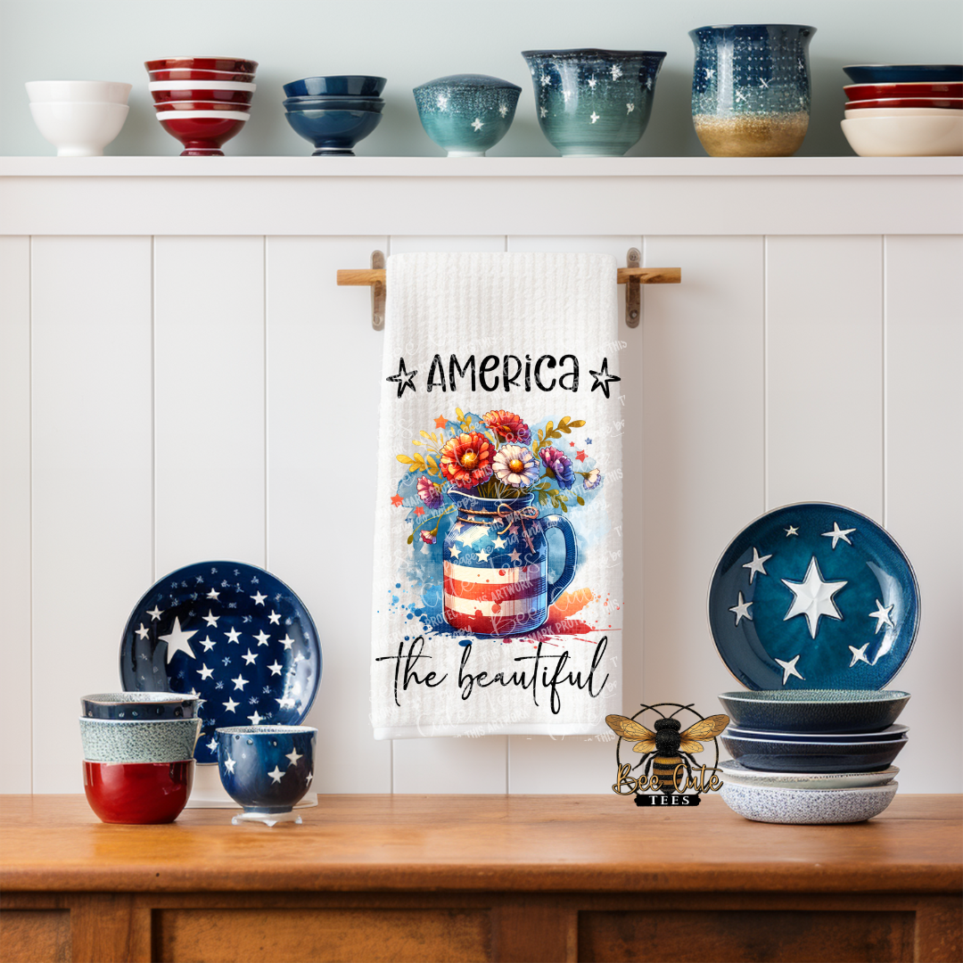 White kitchen towel hanging on a rack in a cozy kitchen, featuring a patriotic mason jar filled with colorful flowers and the phrase “America the Beautiful,” surrounded by star-themed dishes and rustic decor on a wooden counter.
