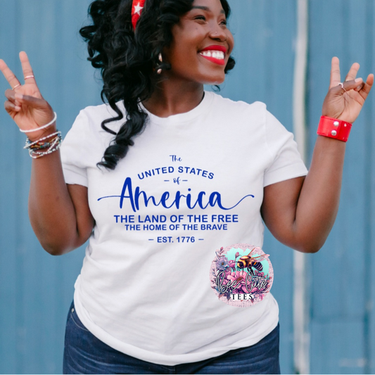 Smiling woman wearing a white graphic tee that reads “The United States of America, The Land of the Free, The Home of the Brave, Est. 1776,” styled with patriotic accessories and posing with peace signs.
