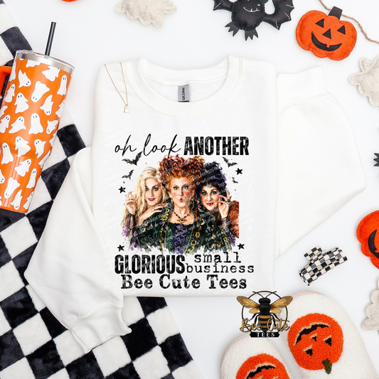 A white crewneck sweatshirt laid flat on a white background with Halloween-themed props. The sweatshirt features a graphic of the three witches with the text 'oh look ANOTHER GLORIOUS small business Bee Cute Tees'.