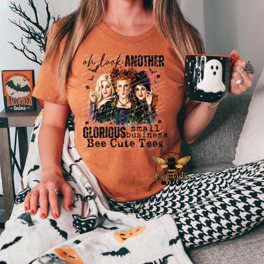 A woman wearing an orange graphic t-shirt with a Halloween-themed print featuring witch characters and the text 'oh look ANOTHER GLORIOUS small business Bee Cute Tees'. She is holding a black mug with a white ghost design, sitting on a gray couch with a black and white houndstooth blanket.