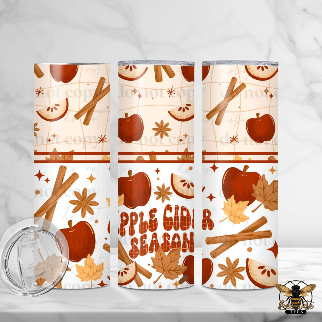 Three insulated tumblers with a fall apple cider season pattern, featuring cinnamon sticks, apples, leaves, and the text 'APPLE CIDER SEASON'. A clear lid is shown in the bottom left on a white marble background.