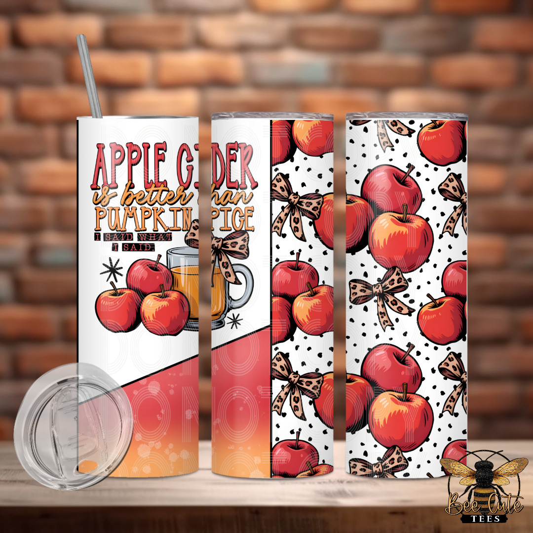 Three views of a skinny tumbler with a wraparound autumn apple design, featuring red apples, brown bows, and polka dots on a white background, with a clear lid and straw shown in front. The tumbler is displayed on a wooden surface with a blurred brick wall background.