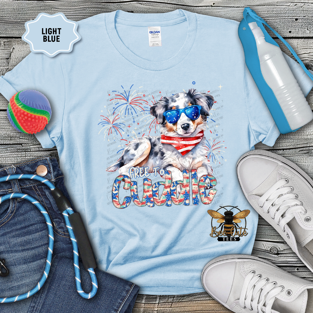 Light blue t-shirt with a dog graphic and text, surrounded by jeans, shoes, and accessories on a wooden surface.