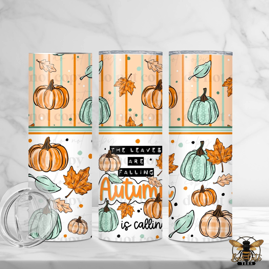Three insulated tumblers with clear lids are shown on a light marble surface. The tumblers feature an autumn-themed wrap design with pumpkins, leaves, stripes, and the phrase 'The Leaves Are Falling Autumn is calling' in black, white, orange, and teal.