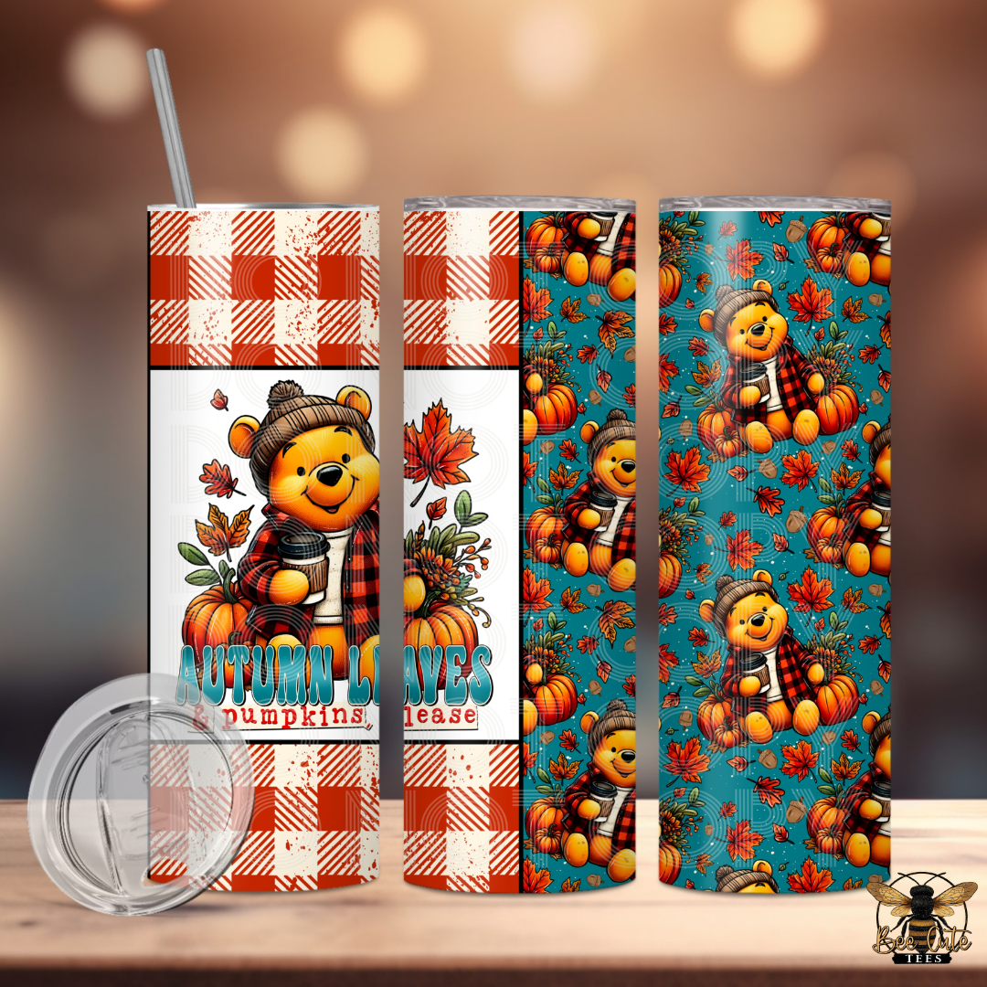 A 20 oz skinny tumbler shown from multiple angles on a light wood surface with a blurred warm bokeh background. The tumbler features an autumn theme with a cartoon bear wearing a beanie and plaid shirt, surrounded by pumpkins, leaves, and the text 'AUTUMN LEAVES & PUMPKINS  PLEASE'.