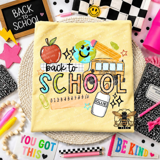 A flat-lay presentation of a pale yellow t-shirt featuring a colorful 'back to school' graphic with an apple, globe, pencil, school bus, glue bottle, and bee illustration. The shirt is styled on a woven mat with school-themed props including notebooks, pens, and a chalkboard.