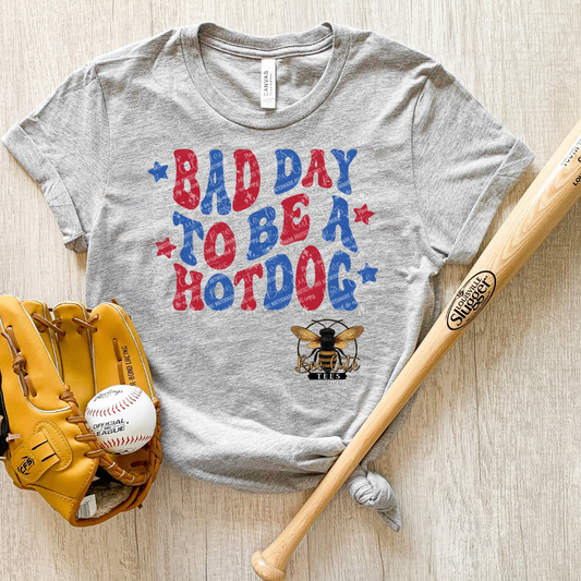 Gray t-shirt with 'Bad Day to Be a Hotdog' text and baseball accessories on a wooden surface