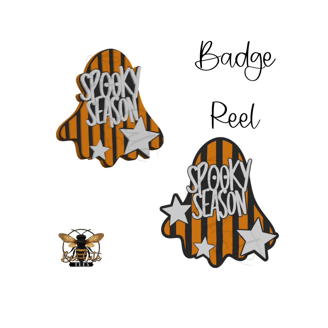 Two ghost-shaped embroidered badges with orange and black vertical stripes, silver glittery stars, and the words 'Spooky Season' in white, shown on a white background. One badge is larger and labeled 'Badge Reel,' and a smaller bee logo appears in the lower left.