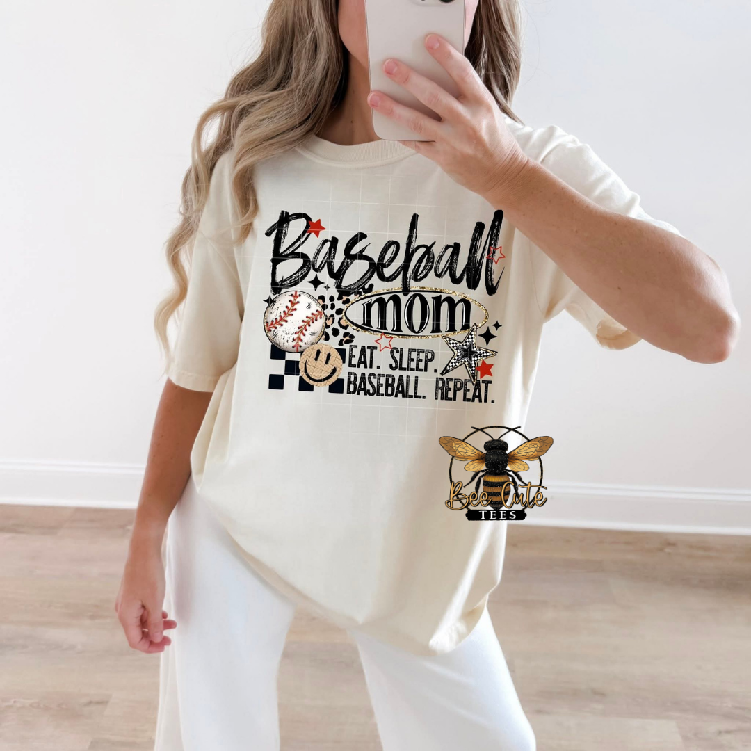 A woman takes a mirror selfie wearing a cream-colored t-shirt with a 'Baseball Mom' graphic print and white pants, set against a neutral indoor background.